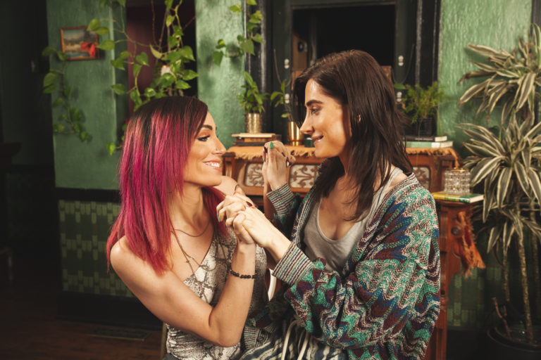 Two transgender women sit close together in a cozy, plant-filled room, smiling warmly and holding hands as they look into each other’s eyes—an intimate, affirming moment of connection and MTF identity exploration.