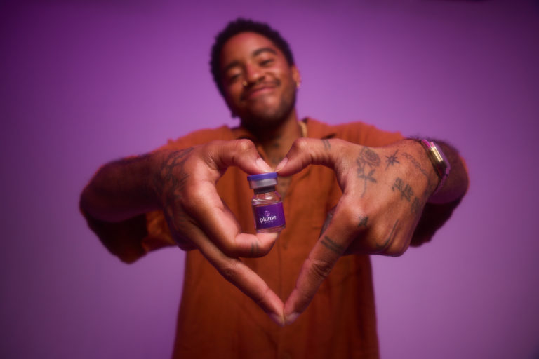 A person forms a heart shape with their hands around a vial of Plume Clinic medication against a purple background, representing gender-affirming care for transgender people.