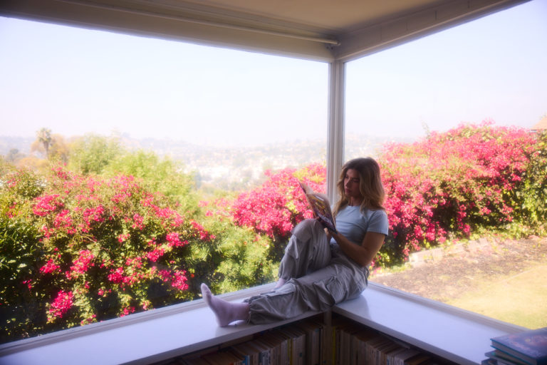 A person sits by a sunny window reading a book, surrounded by blooming pink flowers and greenery, in a lifestyle image related to transgender healthcare.