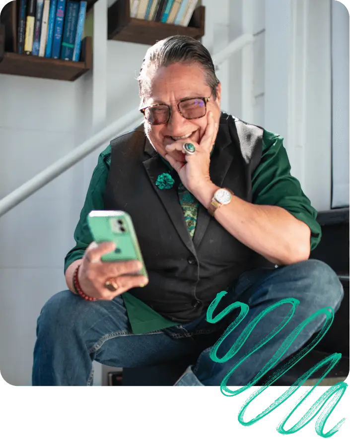 Smiling person wearing glasses and a vest, looking at a smartphone in a bright room with bookshelves in the background.