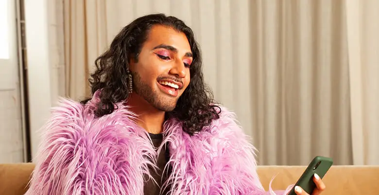 Person smiling while looking at a smartphone, wearing a vibrant pink faux fur coat and bold makeup, seated on a beige couch.