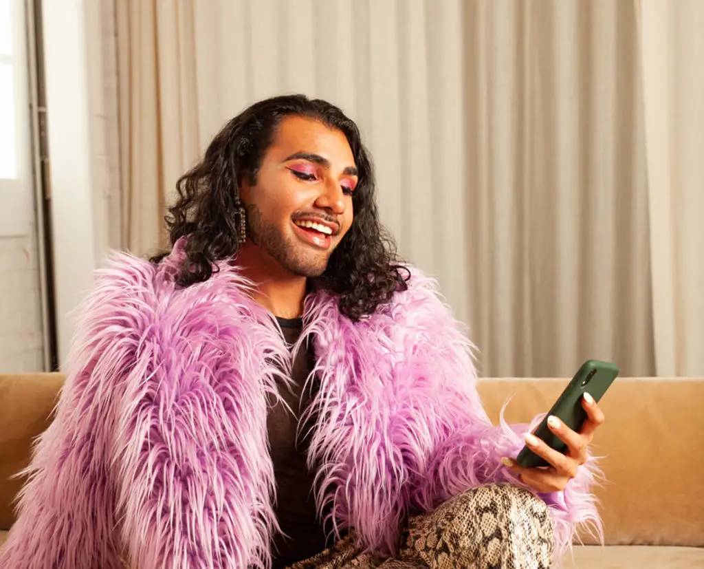 Person smiling while looking at a smartphone, wearing a vibrant pink faux fur coat and bold makeup, seated on a beige couch.