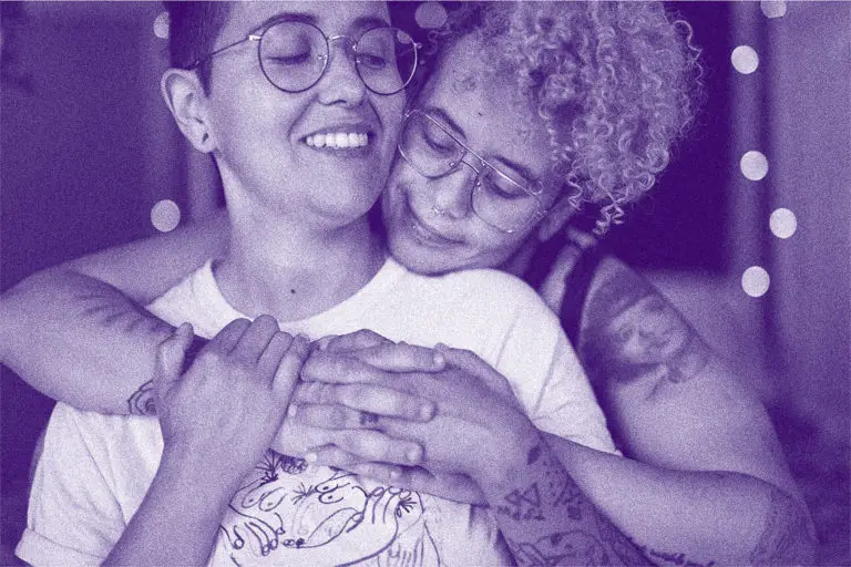 Two transgender friends with a purple overlay. One is wearing a white shirt and glasses. They are being hugged from behind. The person behind them has blonde curly hair.