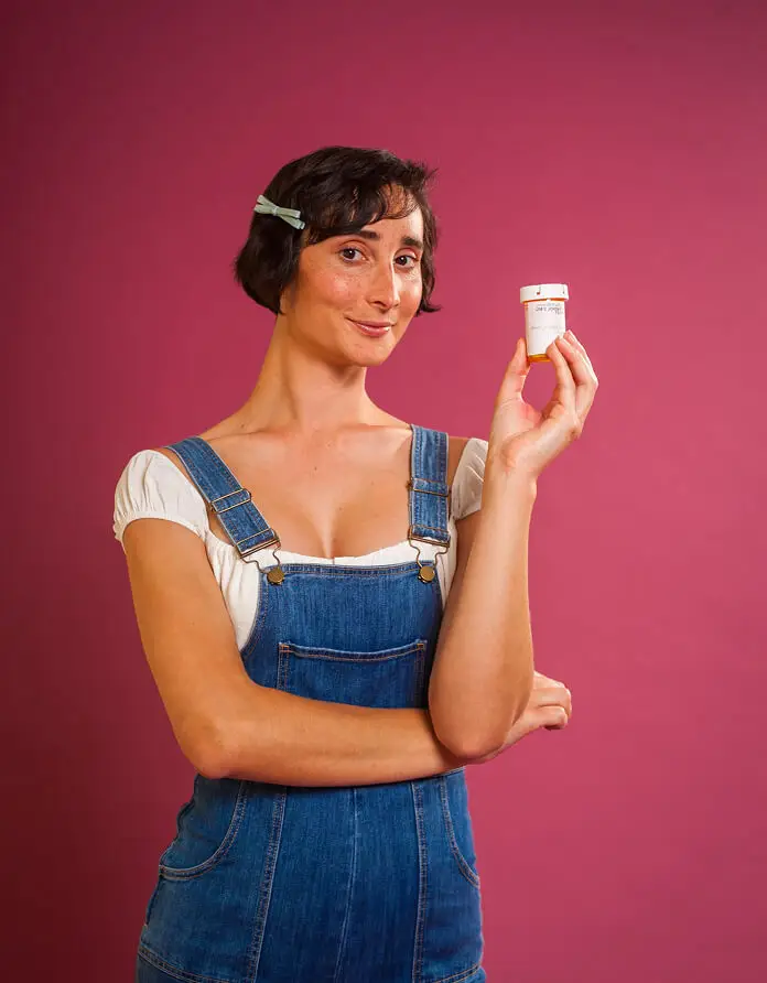 person in denim overalls holding a prescription bottle and smiling confidently, with a dark pink background