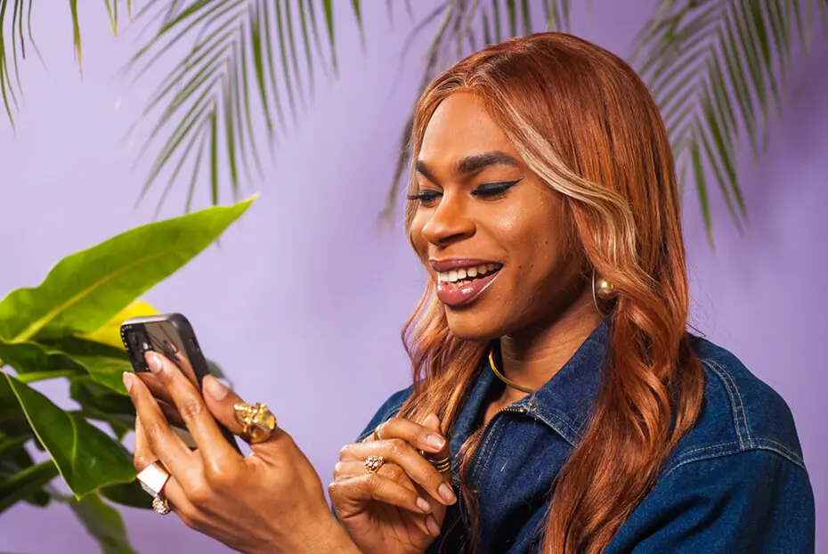 person with long auburn hair smiling while using a smartphone, with tropical plants and a light purple background