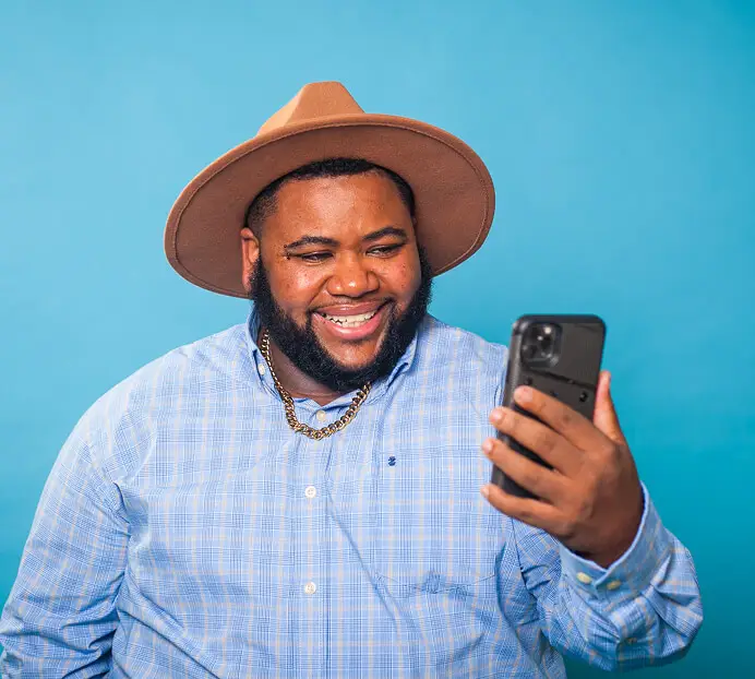 person wearing a brown hat and plaid shirt smiling while looking at a phone, standing against a bright blue background