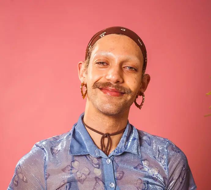 smiling person with heart-shaped earrings and a brown bandana, wearing a sparkly blue shirt, posed against a pink background