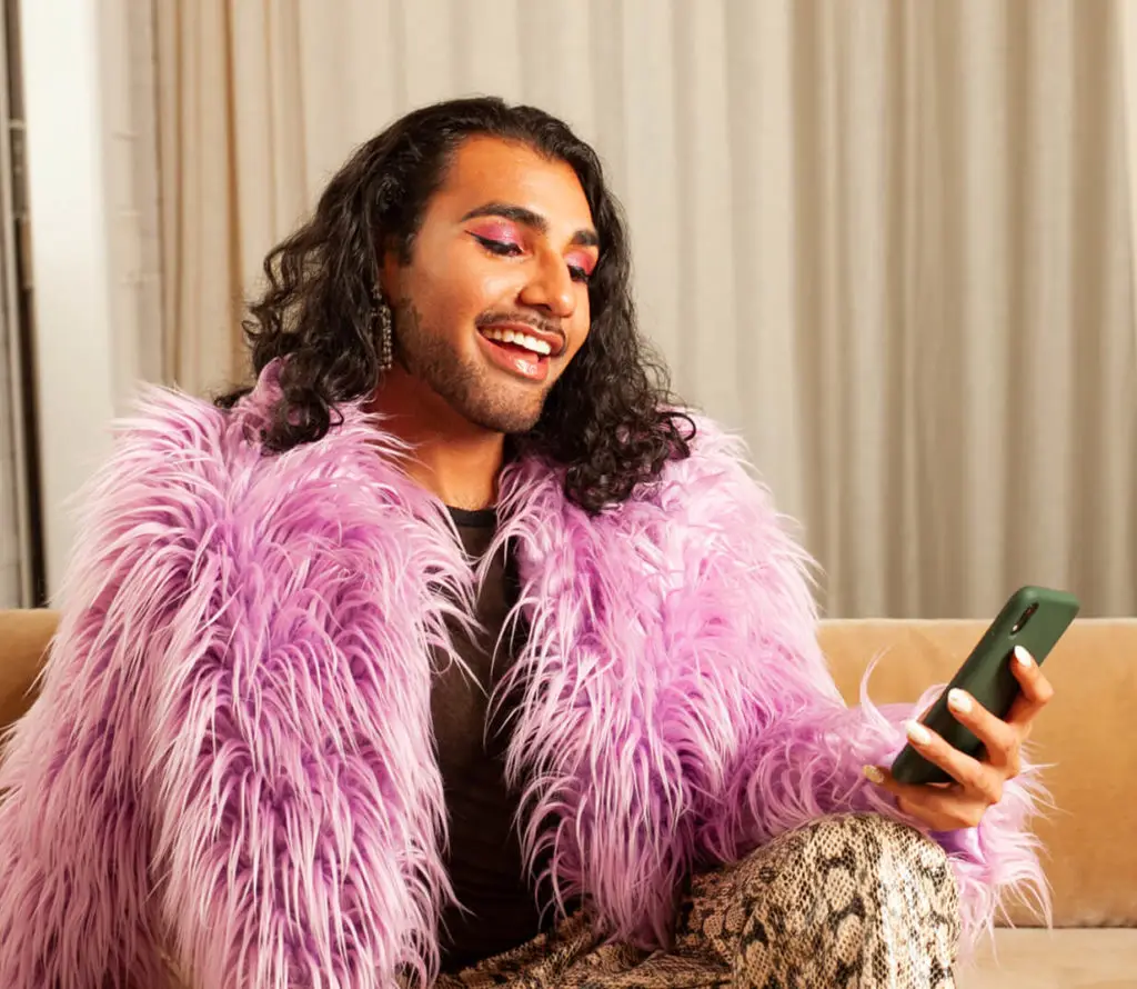 Person smiling while looking at a smartphone, wearing a vibrant pink faux fur coat and bold makeup, seated on a beige couch.