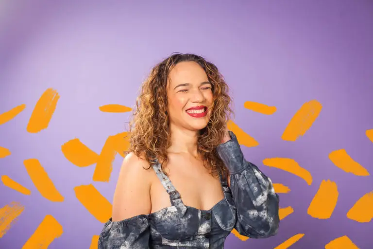 transgender woman smiling in front of a purple background and orange paint marks scattered behind her.