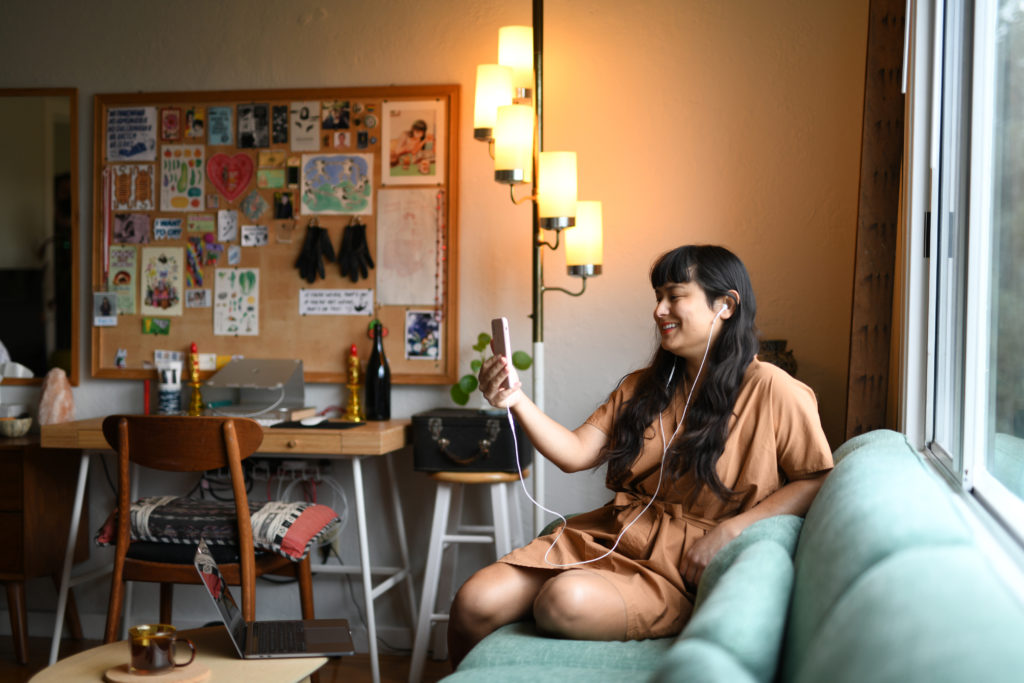 A transgender woman in a brown dress sits on a green couch near a window, smiling at her phone with earphones in during her trans telehealth appointment for gender-affirming hormone therapy (HRT). A bright, cozy room with a decorated bulletin board and a lit floor lamp in the background creates a relaxed atmosphere.