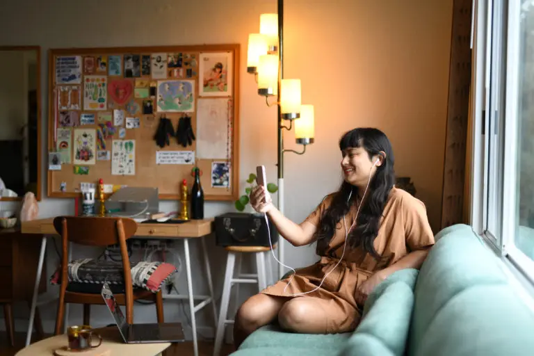 A transgender woman in a brown dress sits on a green couch near a window, smiling at her phone with earphones in during her trans telehealth appointment for gender-affirming hormone therapy (HRT). A bright, cozy room with a decorated bulletin board and a lit floor lamp in the background creates a relaxed atmosphere.