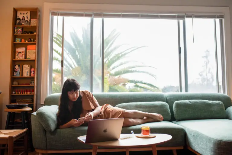 A woman in a brown dress lounging on a green couch, using a laptop while holding a phone. Cover photo for the article Trans Healthcare Resources in Florida.