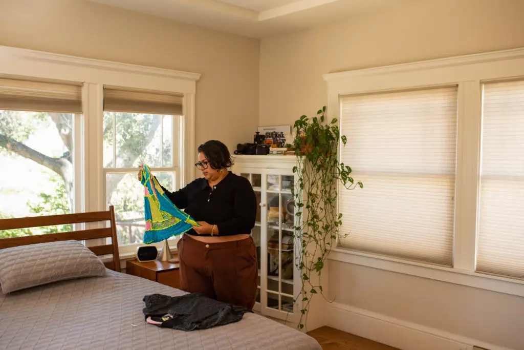 A transgender person moving into their new home. They are standing in a cozy bedroom, examining a colorful piece of fabric. Large windows with natural light illuminate the room, which has a bed, a cabinet, and plants, creating a warm and relaxed atmosphere.