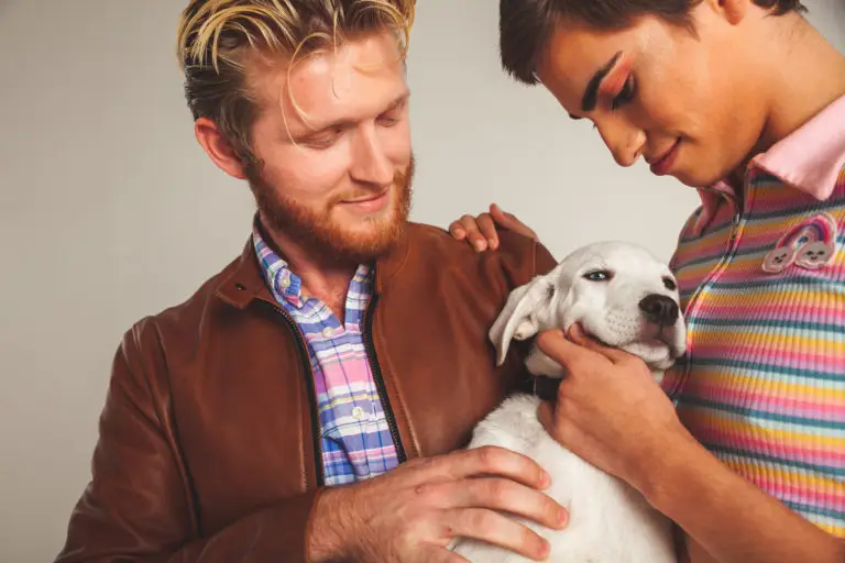 Two people stand close together, gently holding and petting a white puppy—a simple moment that subtly reminds us how connection and compassion can help ease mental health disparities. The person on the left wears a brown jacket; the other, a colorful striped top.