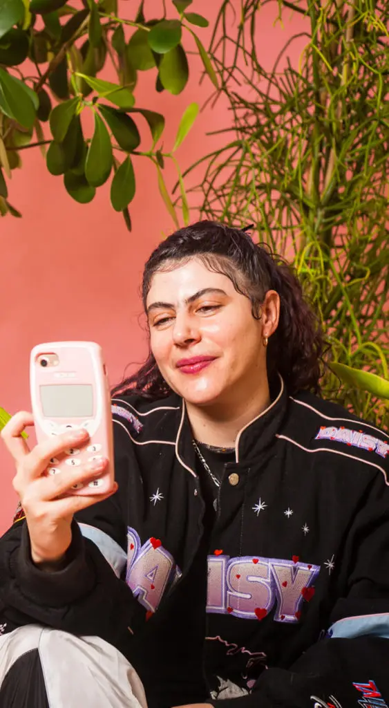 A non-binary person sitting amongst plants, using their phone