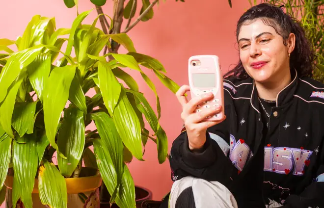 A non-binary person sitting amongst plants, using their phone