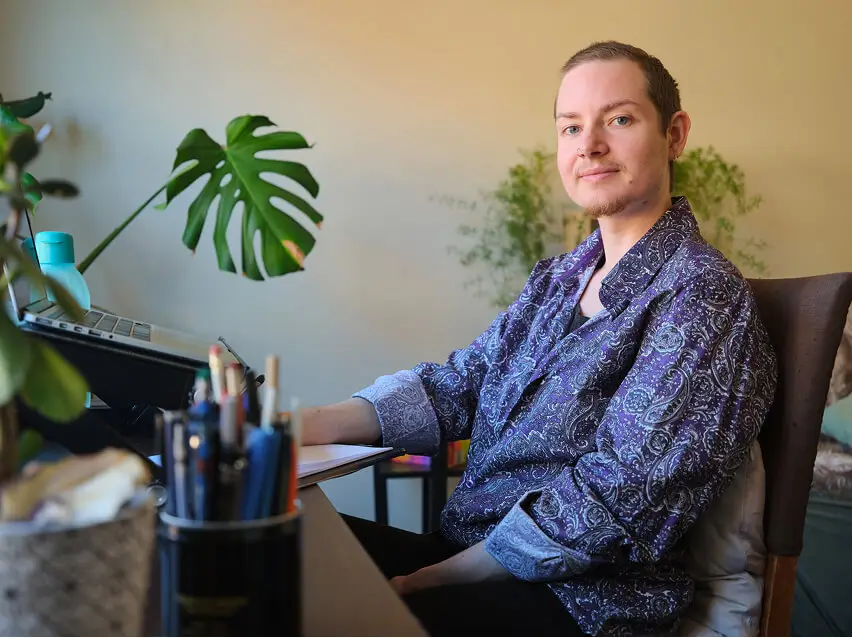 Person smiling sitting at a desk in a home office, surrounded by plants and working on a laptop