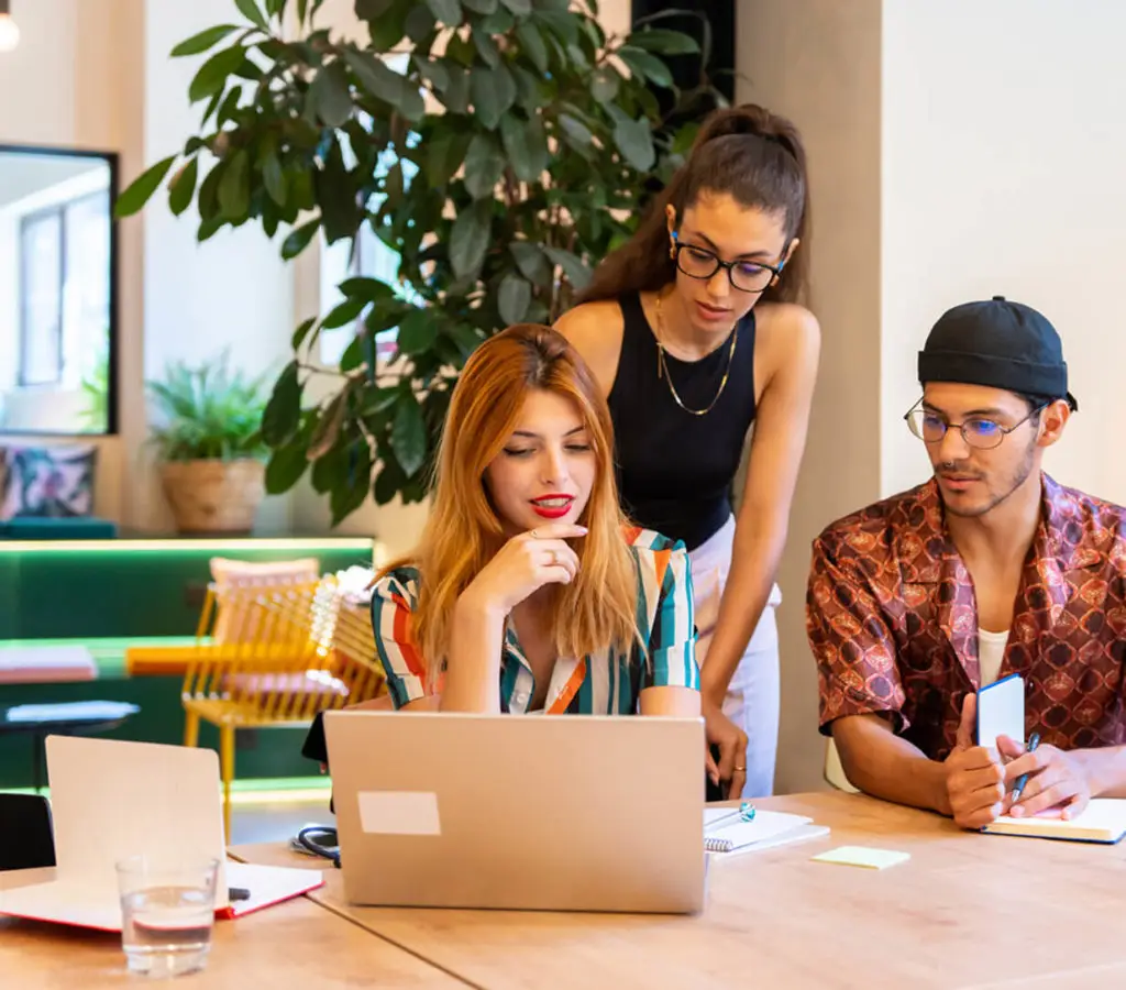 Three coworkers collaborating at a table with a laptop and notebooks, in a modern, plant-filled coworking space
