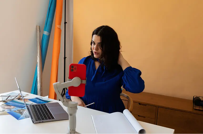 Person with long dark hair speaking on a livestream with a phone and laptop, seated at a colorful creative workspace