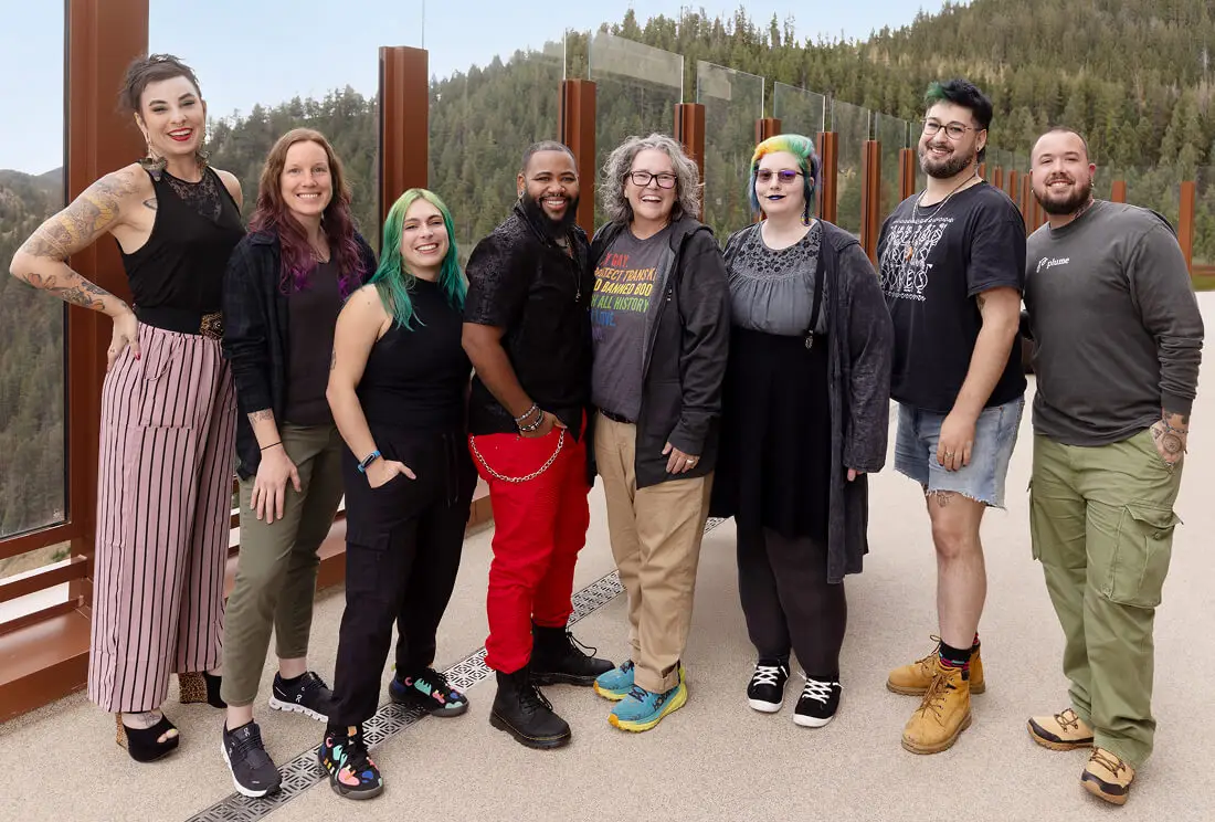 A group of proud trans-health experts in plainclothes standing side-by-side in front of a tree-covered mountain in the background