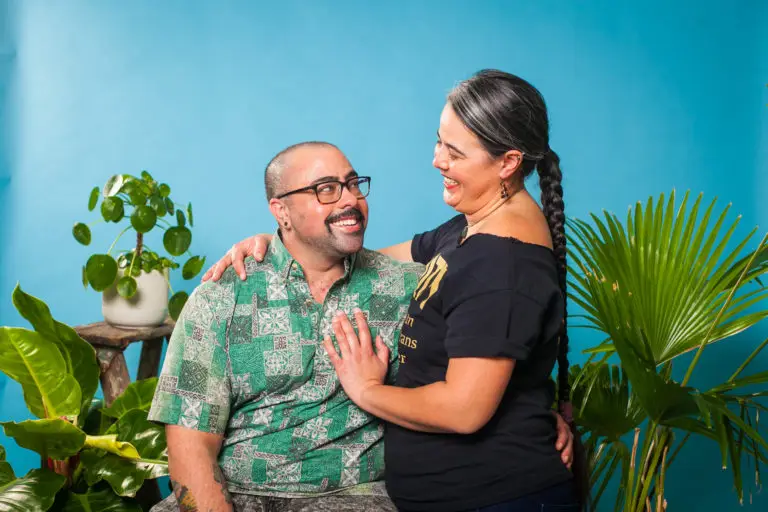 A smiling man sits while a woman stands beside him with her hand on his chest. Both are looking at each other, surrounded by green plants against a blue background, celebrating love and connection within the trans Hispanics community.