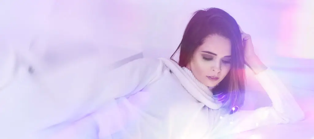 person in white turtleneck lying down in soft rainbow light on a bed