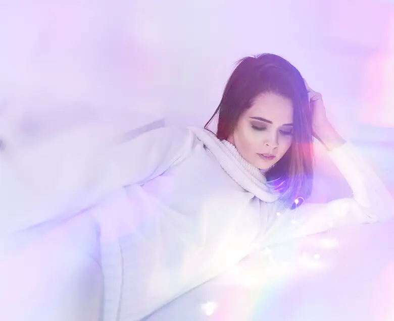 person in white turtleneck lying down in soft rainbow light on a bed