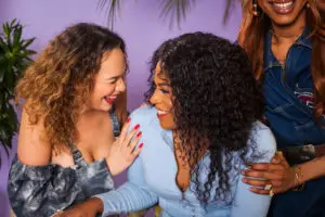 Three transfeminine friends smiling and supporting one another, sharing a joyful moment of sisterhood and care — a visual echo of “protect the dolls,” honoring love and safety within trans femme community.