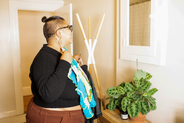 Person smiling in front of a mirror while holding colorful clothing choices, representing self-expression, confidence, and gender-affirming care as part of transgender and nonbinary identity.