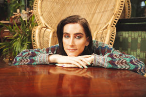 Transgender person with short dark hair and subtle makeup resting their chin on folded hands at a wooden table, seated in front of a wicker chair in a warm, plant-filled room.