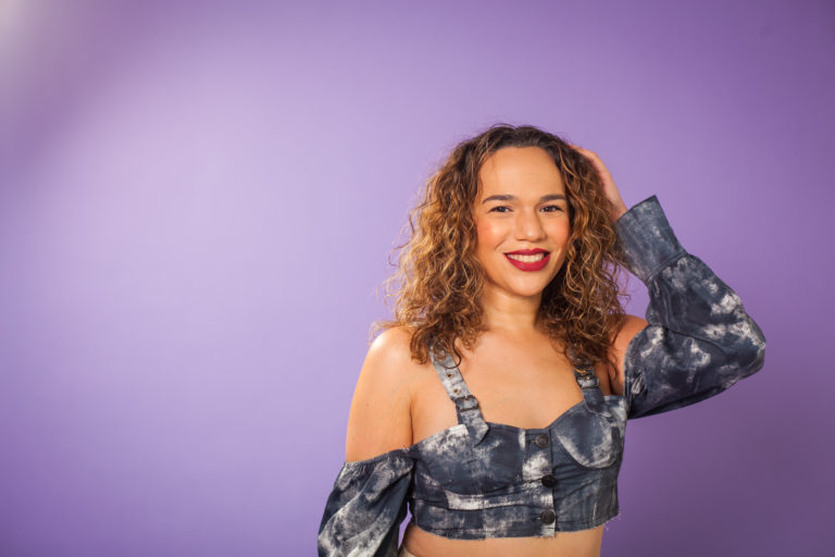 A full-color portrait of a smiling transgender woman with curly, light brown hair, posed against a solid purple studio background. She is wearing a distinctive blue denim tie-dye crop top with matching, partially donned sleeves, her left hand placed behind her head.