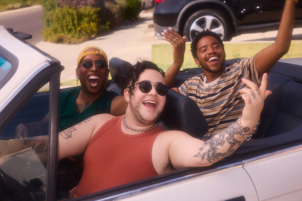 A joyful group of three transgender individuals celebrate pride while riding in a white convertible car on a sunny day. In the foreground, a person with curly hair, sunglasses, and an orange tank top smiles widely, gesturing out the window and showcasing arm tattoos. In the back, two friends laugh heartily—one wearing a yellow cap and green top, and the other in a striped shirt with an arm raised high in celebration.