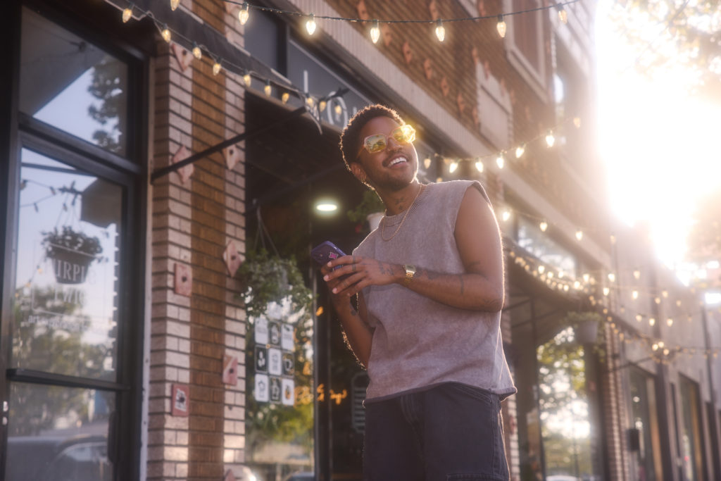 A joyful transgender individual stands on a city sidewalk, radiating pride during a sun-drenched golden hour. Bathed in warm, glowing light that creates a bright lens flare, they smile broadly while wearing yellow-tinted sunglasses and a grey sleeveless knit top, holding a smartphone beneath strings of festive overhead market lights.