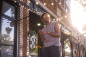 A joyful transgender individual stands on a city sidewalk, radiating pride during a sun-drenched golden hour. Bathed in warm, glowing light that creates a bright lens flare, they smile broadly while wearing yellow-tinted sunglasses and a grey sleeveless knit top, holding a smartphone beneath strings of festive overhead market lights.