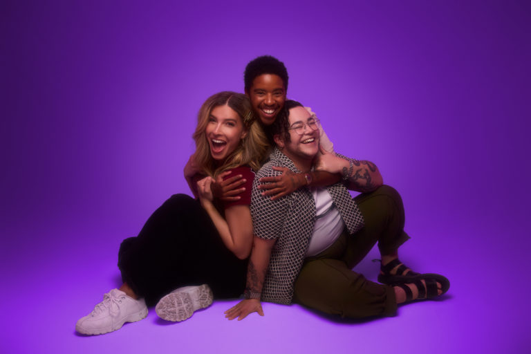 Three smiling transgender adults pose together against a purple studio backdrop. One person hugs the other two from behind as they sit close together, laughing and embracing, conveying joy, friendship, and community.