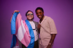 Two transgender adults smiling and holding a transgender pride flag, representing community and visibility in a guide about voting as a transgender person in 2026.