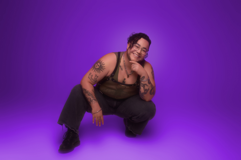 A smiling transgender person with tattoos and glasses crouches against a vibrant purple studio backdrop.